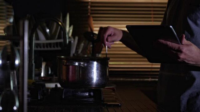 A mid-shot captures a focused and modern mood as a man stirs a pot on a gas stove while referencing a digital tablet, suggesting technology-assisted home cooking in a dimly lit kitchen