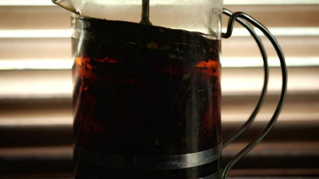 A close-up shot captures a warm, intimate mood as a French press, filled with brewing tea or coffee, is silhouetted against the soft light filtering through horizontal window blinds