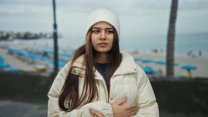 Woman in white coat and beanie stands by a seaside with palm trees and umbrellas, looking cold and...