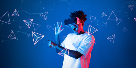 Smart boy standing at blue background while floating geometric shape. African man playing game and wearing VR goggle and casual shirt. Augmented reality technology innovation concept. Ingenuity.