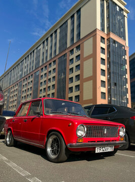 A classic custom tuned red Lada VAZ 2101 Zhiguli car with stylish alloy wheels parked outside a modern residential apartment building.