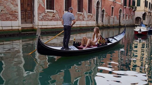 Experienced gondolier expertly navigating traditional wooden gondola with female tourist through scenic Venice canal, capturing serene Venetian urban landscape