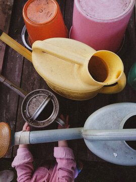 Colorful Gardening Tools and Child's Hands on Wooden Deck &ndash; Top View. Rustic gardening scene featuring colorful plastic buckets and watering cans arranged on an aged wooden deck.