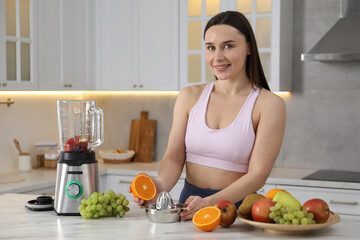 Woman in fitness clothes with orange and juicer making juice at white marble table indoors