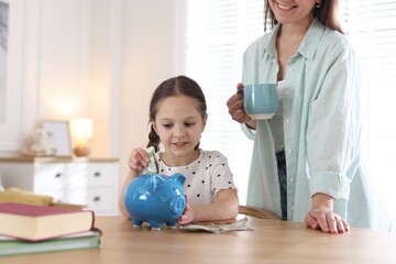 Fototapeta premium Mother and daughter putting pocket money into piggybank at table indoors, closeup