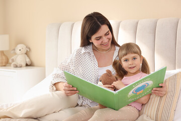 Bedtime. Happy little daughter reading book with her mother on bed at home