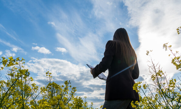 A person stands amid vibrant yellow flowers, holding a digital tablet and gazing up at the expansive sky filled with clouds, enjoying a sunny day outdoors