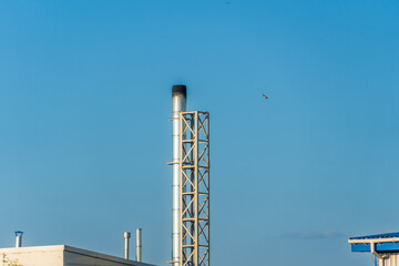 A tall smokestack rises against a bright blue sky, with wisps of smoke billowing. A small bird can be seen soaring in the distance during midday