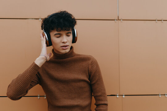 Fashionable young man standing in front of an orange wall, wearing headphones and listening to music with hands on head - Powered by Adobe