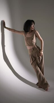 Vertical video: Model posing in stylish beige outfit, leaning against wall in fashion studio