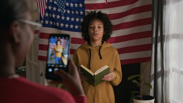 Rack focus over the shoulder shot of mother taking photos of cheerful pre-teen son holding book with green cover and reading out patriotic poem standing against American flag hanging in living room