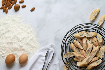 View from above of a white marble slab with ingredients for Italian pastries. Flour, eggs and almonds. Cantuccini, which are already baked, lie next to them in a metal basket.