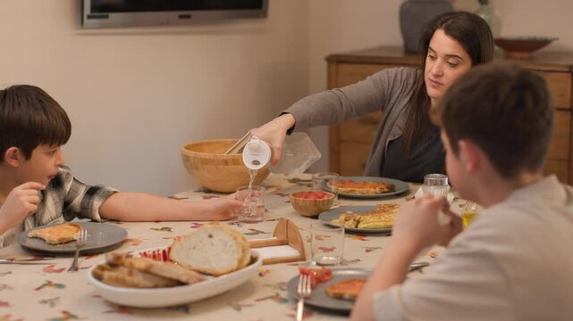 Medium shot of a mother and two children seated at the home dining table, ready to eat. She pours water as they prepare to enjoy a healthy Mediterranean meal with omelet, salad, bread. Horizontal, 4K