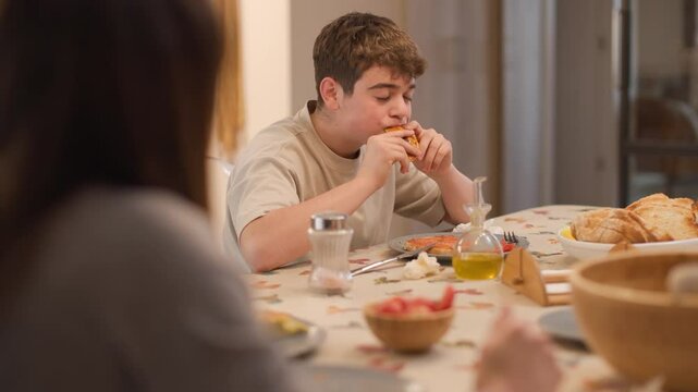 Medium shot of a boy reaching for an empanada from the central plate on the kitchen table during a family meal. The mother stands in the background. Homemade food, relaxed and hungry moment.Horizontal