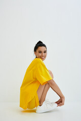 Smiling woman wearing a bright yellow oversized t-shirt and white sneakers, sitting in a relaxed pose against a plain white background with natural lighting.