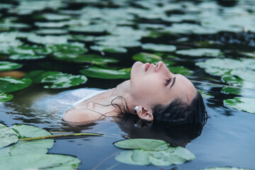 Sensual portrait of young beautiful brunette woman posing lying in river in water lilies