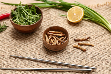 Wooden bowl with fried maggots, arugula, lemon and green onion on beige background