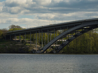 Obraz premium Highway bridge spanning river, concrete pillars rising through verdant landscape, connecting terrain beneath overcast sky, representing contemporary transportation infrastructure