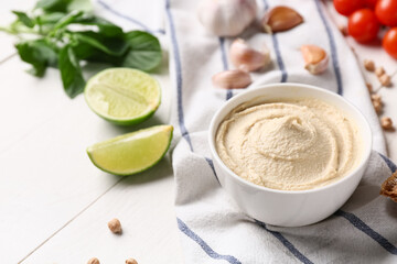 Bowl of tasty hummus with ingredients and napkin on white wooden background, closeup