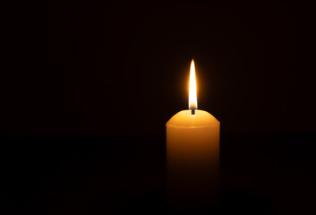 A single burning candle flame or light glowing on a big white candle isolated on black or dark...