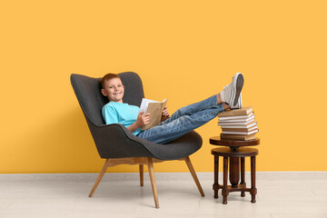 Cute little boy reading book while sitting in armchair near color wall