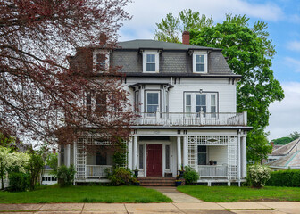 Elegant two-story house with mansard roof and dormer windows surrounded by trees and shrubs in...