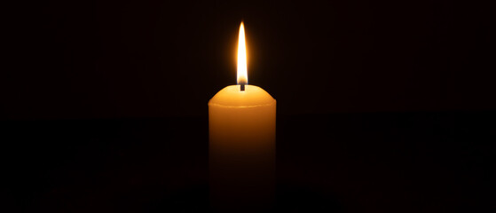 A single burning candle flame or light glowing on a big white candle isolated on black or dark...