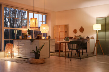 Interior of modern kitchen with counters, dining table and glowing lamps in evening