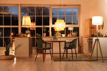 Interior of modern kitchen with counters, dining table and glowing lamps in evening