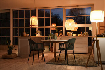 Interior of modern kitchen with counters, dining table and glowing lamps in evening