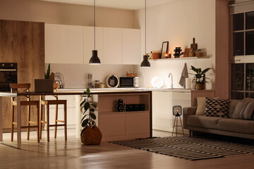 Interior of modern kitchen with counters, table and glowing lamps at night