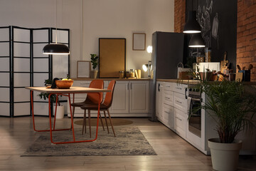 Interior of stylish kitchen with counters, dining table and glowing lamps at night