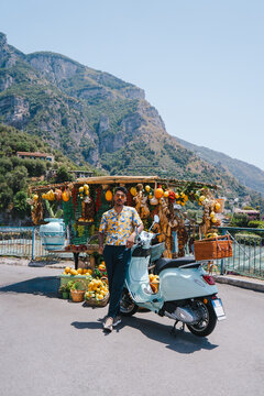 Man standing next to vintage scooter and colorful fruit stand on the Amalfi Coast, Italy..