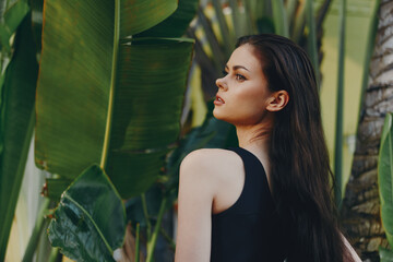 Attractive woman with long black hair standing near lush green plants and palm trees on sunny day