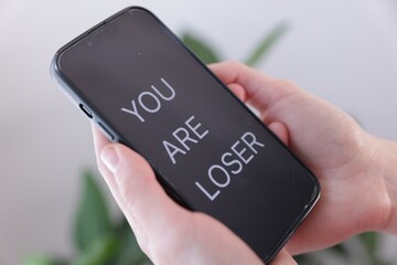 Cyber bullying. Woman holding smartphone with text You Are Loser on blurred background, closeup