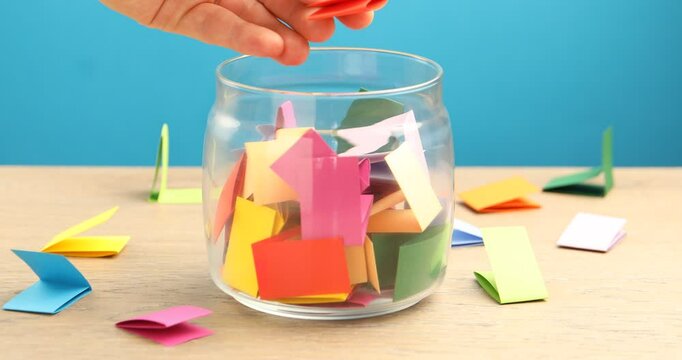 Woman taking color paper piece form glass jar at wooden table against light blue background, closeup
