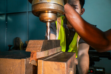 An industrial worker operating a drill press in a workshop. The focus is on the precision and concentration required to handle machinery. The worker wears a safety vest, ensuring a secure work.