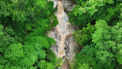 Drone aerial view shows a swollen muddy river rushing through dense forest, highlighting the impact of flash flooding as water surges over rocks, causing rapid and turbulent flow in natural terrain.
