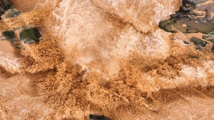 Bird’s eye perspective captures sediment-rich floodwaters rushing over a barrier, highlighting how extreme weather and global warming amplify natural flood risks. Thailand.
