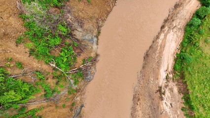 Overhead shot reveals a muddy riverbank eroded by a recent landslide, emphasizing the role of deforestation and heavy rain in destabilizing slopes and increasing flood risks.
