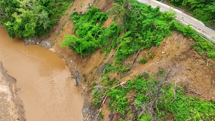 Drone aerial view shows a landslide on a deforested slope next to a muddy river, illustrating how heavy rains and loss of vegetation accelerate soil erosion and slope failures. Thailand.
