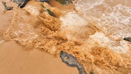 Bird’s eye perspective shows turbulent sediment-rich floodwaters surging through rocks, illustrating the dynamic movement and erosion caused by flooding. Thailand.
