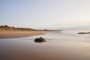Calm expansive beach with soft waves, scattered rocks, distant sand dunes, and gentle morning light creating peaceful atmosphere