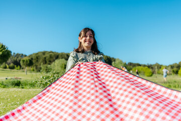 Smiling girl holding red and white checkered picnic blanket in park on sunny day, ready for outdoor meal