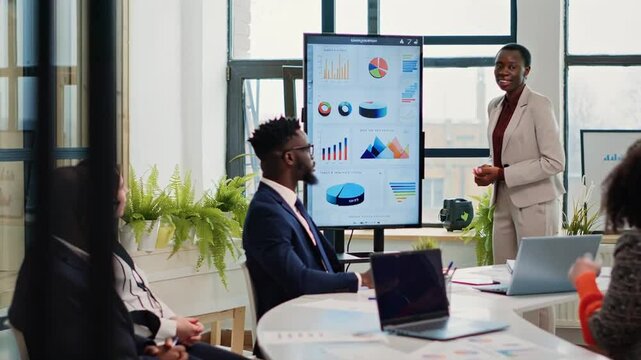 Black businesswoman showing charts on digital screen while presenting project to colleagues in modern office meeting room, explaining graphs and discussing financial results and corporate strategy