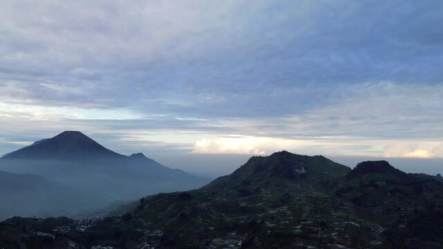 A wide shot of a mountain range under a cloudy sky. The sun is rising or setting, casting a soft light on the landscape. There is a small town in the valley.