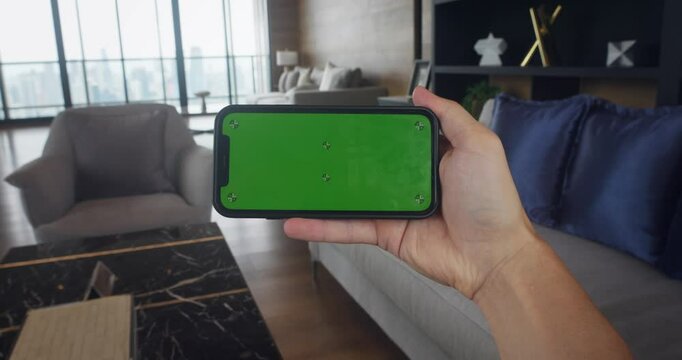 Smart phone with green chroma key mock up screen in with horizontal position at home background. Male hand hold smart phone with chroma key green screen at living room