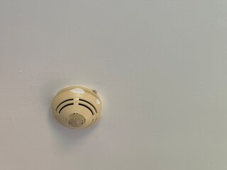 The image shows an off-white smoke detector mounted on a white ceiling, appearing to be an older model due to some discoloration and wear around its edges