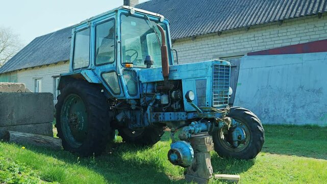 Blue Soviet tractor missing wheel in bright sun beside rural farm building