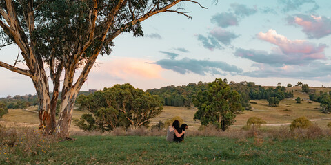 A woman and her dog share a quiet, heartfelt moment under a towering gum tree, gazing out over the rolling hills and colorful evening sky